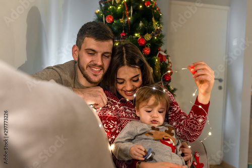 Happy parents with baby in decorated room for Christmas. Young family with son at Christmas tree at home. Happy couple with baby celebrating Christmas together at home