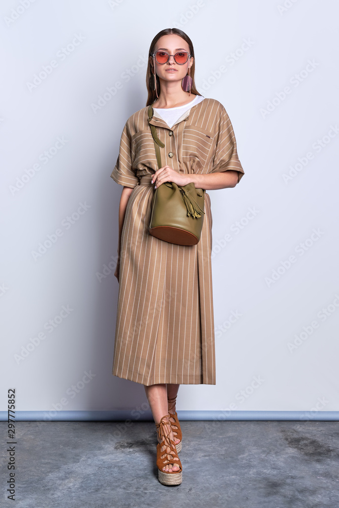 Trendy lady in big sunglasses wearing striped olive dress with leather handbag posing over gray background