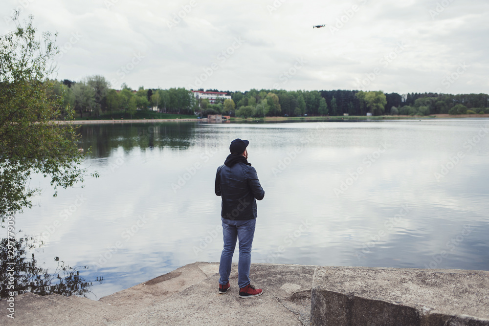 a man in a leather jacket controls a drone in the background of the lake