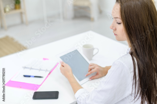 A young woman in a white shirt and jeans sits at a table with a tablet in her hands. A female office manager performs work on a modern device. The view from behind.