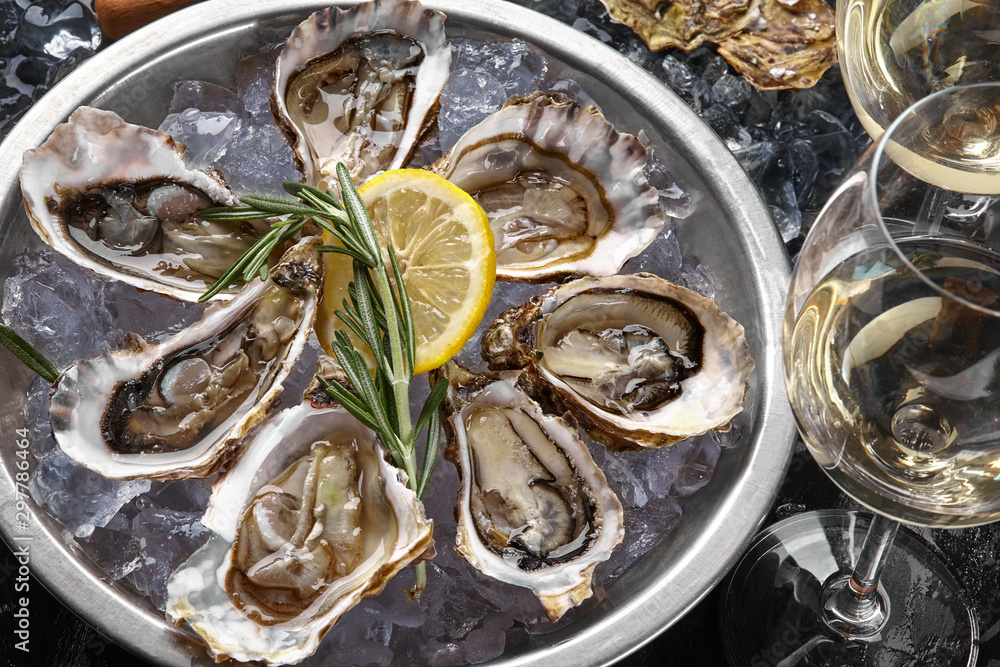 Fresh opened oysters, ice, lemon on a round metal plate and champagne are on a black stone textured background. Top view. Close-up shot.