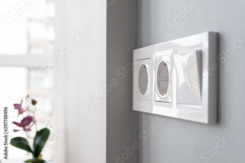 A closeup view of a group of white european electrical outlets and a switch located on a gray wall in a light modern kitchen by the window. Selective focus. Blurred background photo