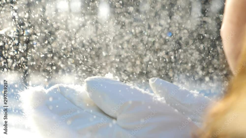 Unrecognizable woman in white gloves blowing off snow from her palms. Young girl standing among winter forest and playing with snow at sunny day. Blurred background. Winter concept Slow mo Detail view
