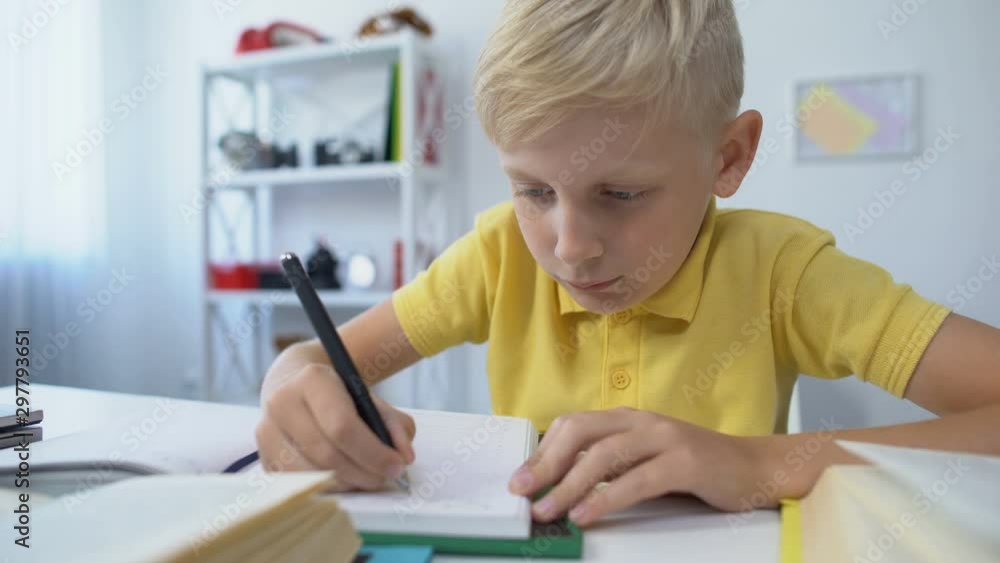 Blond male kid writing hometask in notebook and putting head on table, boredom Blond male kid writing hometask in notebook and putting head on table, boredom