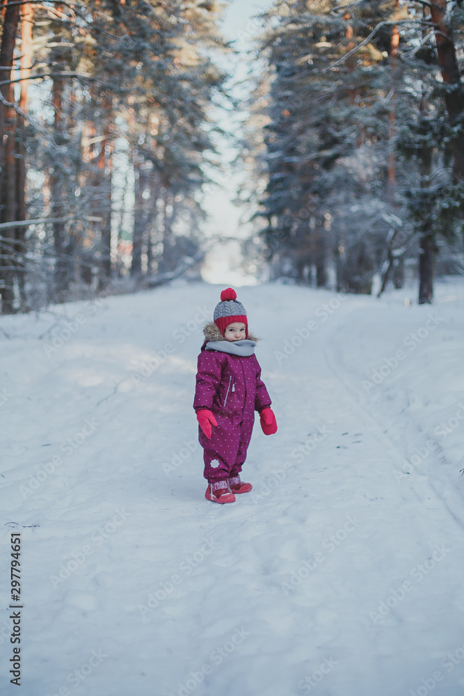 little cute girl 2 years old in lilac overalls walks in a winter snowy forest