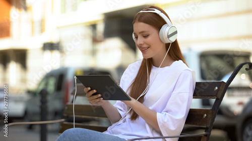 Happy red-haired girl in headset listening to music on tablet outdoor, rest time