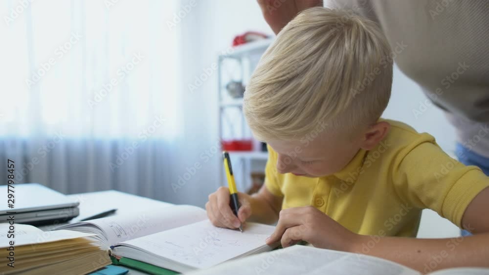 Diligent male kid doing homework at desk, mother praising son, touching head Diligent male kid doing homework at desk, mother praising son, touching head