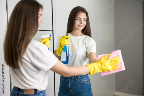 beautiful young smiling woman in yellow gloves with detergent spray in her hand wiping mirror glass with a rag