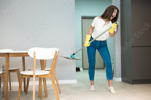cute young woman in yellow gloves sings holding mop as a microphone while washing kitchen floor plays rock star