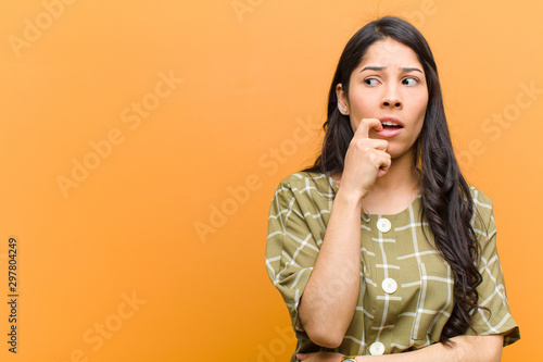 young pretty hispanic woman with surprised, nervous, worried or frightened look, looking to the side towards copy space against brown wall photo