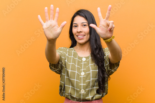 young pretty hispanic woman smiling and looking friendly, showing number eight or eighth with hand forward, counting down against brown wall photo