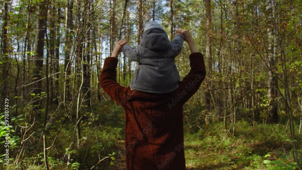 Baby sits on shoulders mother, they walk in autumn forest. Shot behind Baby sits on shoulders mother, they walk in autumn forest. Shot behind