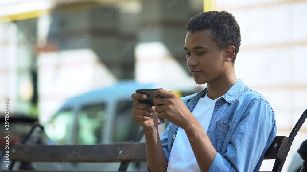 Excited teenager playing action game on smartphone sitting on bench, addiction Excited teenager playing action game on smartphone sitting on bench, addiction