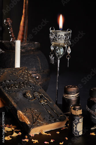 Fototapeta Naklejka Na Ścianę i Meble -  Books with spells, old pot, jars of potion are on a wooden dark table. Petals of dried roses are scattered nearby. Black background. Close-up shot.