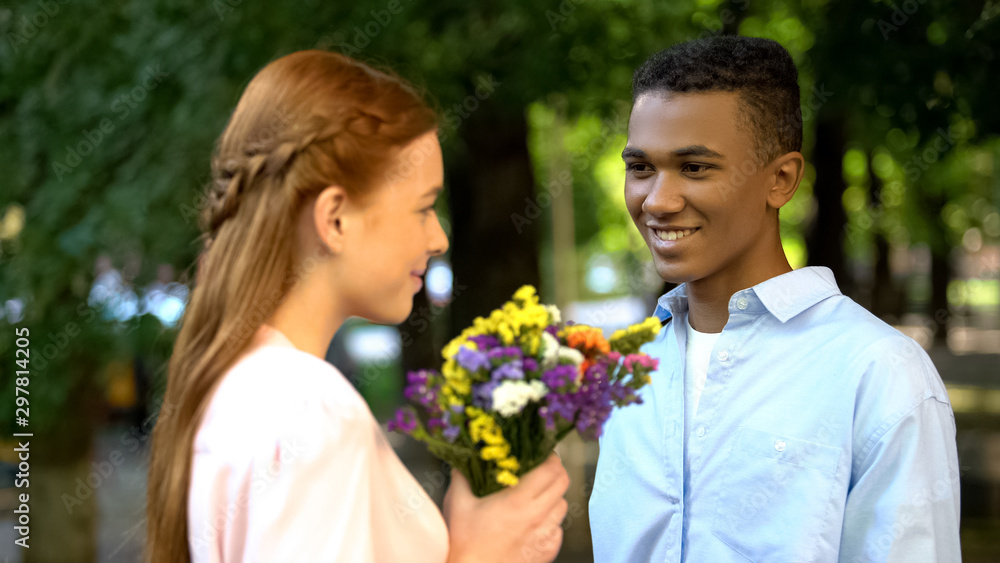 Sweet Afro-American teen boy presenting bunch of flowers to shy girlfriend