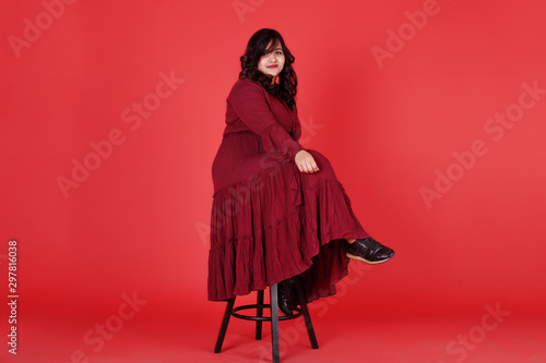 Attractive south asian woman in deep red gown dress posed at studio on pink background sitting on chair.