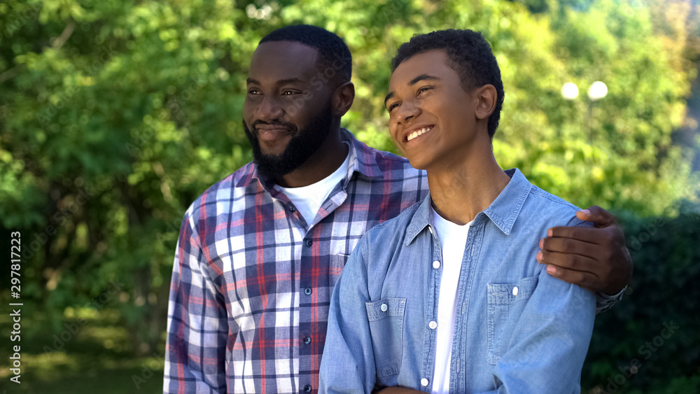 Happy afro-american dad hugging teenage son standing outdoors together, love Happy afro-american dad hugging teenage son standing outdoors together, love