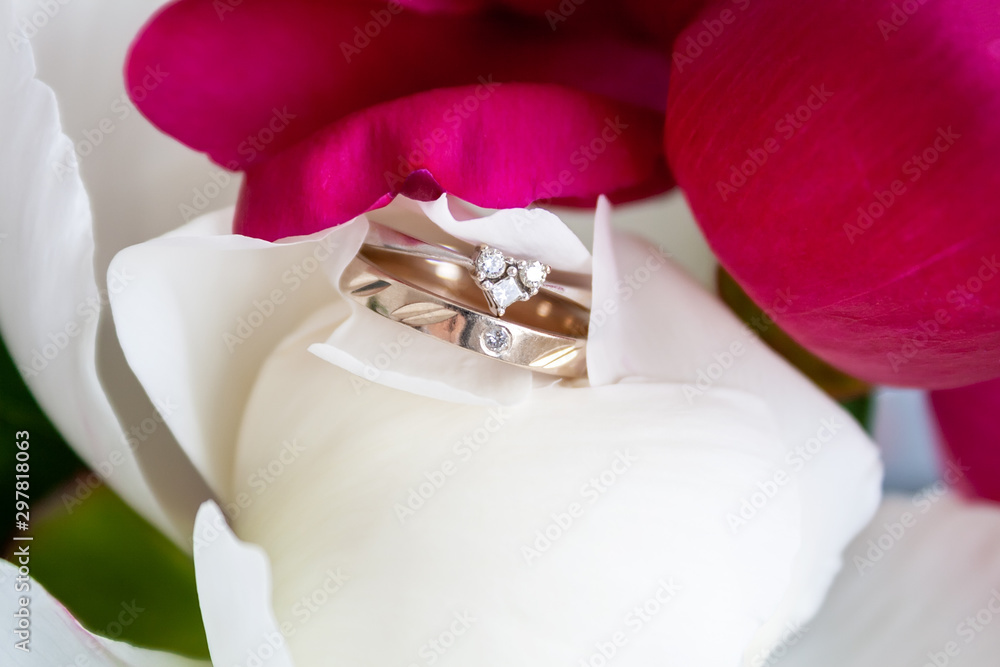 A closeup view of a two beautiful white gold rings: engagement with three small diamonds in the shape of a heart and wedding ring with one small diamond, which lie among the petals of white peony bud A closeup view of a two beautiful white gold rings: engagement with three small diamonds in the shape of a heart and wedding ring with one small diamond, which lie among the petals of white peony bud