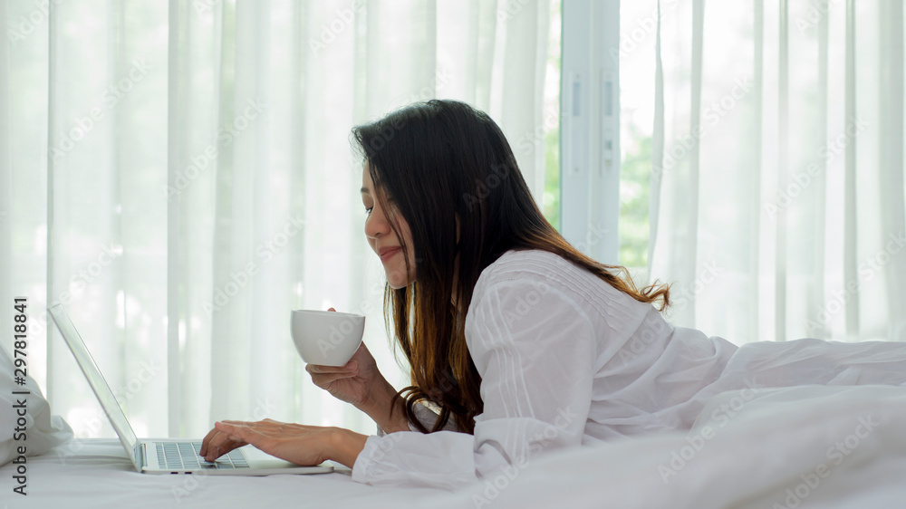 Relax Asian woman lay down in bedroom holding coffee cup using laptop computer Relax Asian woman lay down in bedroom holding coffee cup using laptop computer