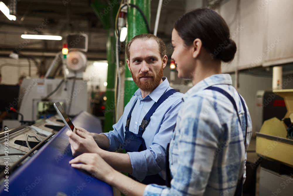 Two engineers in overalls using digital tablet and talking to each other during their work in the plant Two engineers in overalls using digital tablet and talking to each other during their work in the plant