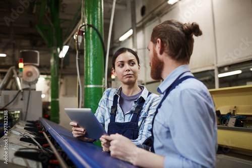Young female worker holding digital tablet and talking to her colleague while they standing near the machine in the plant