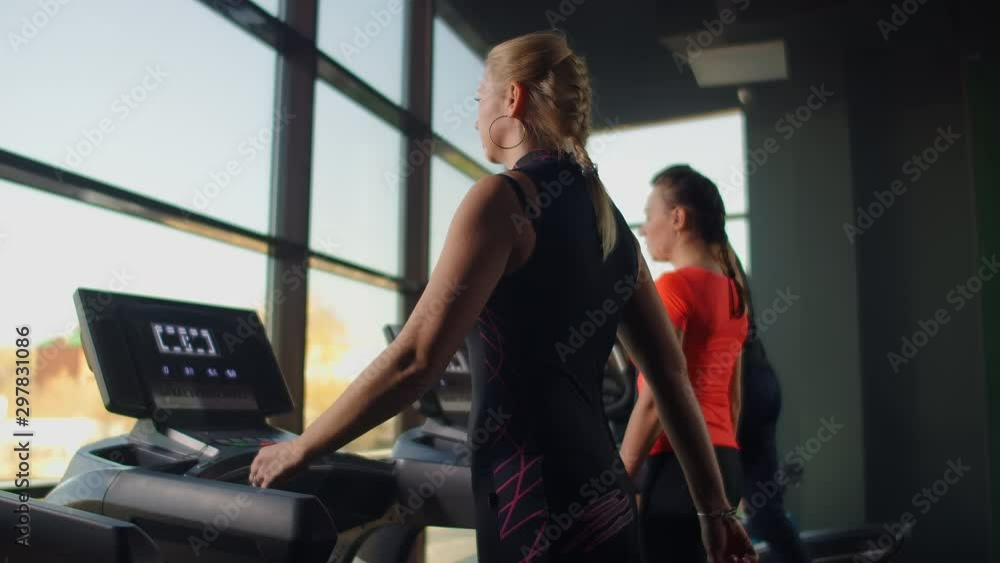 A young beautiful girl walks on a treadmill in front of a large panoramic window in slow motion, against the background of several people A young beautiful girl walks on a treadmill in front of a large panoramic window in slow motion, against the background of several people