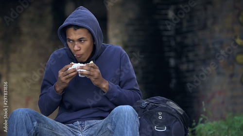 Vászonkép Starving troubled teen chewing snack sitting outdoors, dangerous city suburb