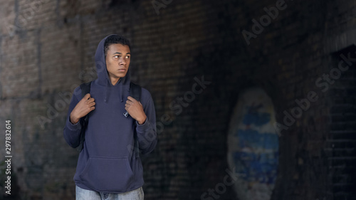 Thoughtful black teen in hoodie with backpack walking dangerous city suburb © motortion