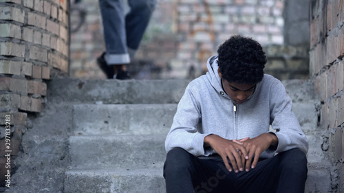 Sad black teenager sitting on stairs in gateway, problem of bullying at school