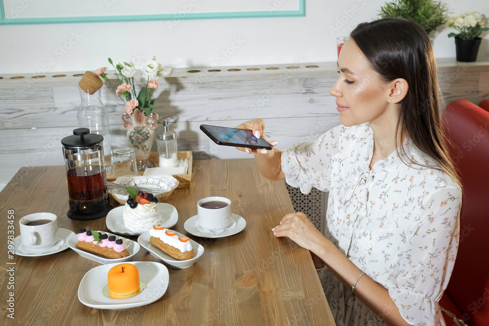 smiling dark haired girl takes picture of tasty natural cakes and tea cups with smartphone at table in cafe, side view smiling dark haired girl takes picture of tasty natural cakes and tea cups with smartphone at table in cafe, side view