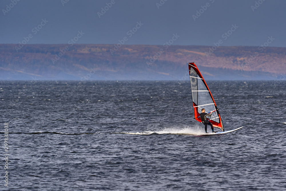 A male athlete is interested in windsurfing. He moves on a Sailboard on a large lake on an autumn day. A male athlete is interested in windsurfing. He moves on a Sailboard on a large lake on an autumn day.