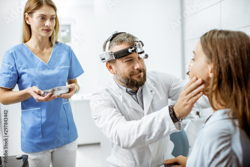 Patient with senior otolaryngologist and female assistant in ENT office during a medical examination, doctor examining nose photo
