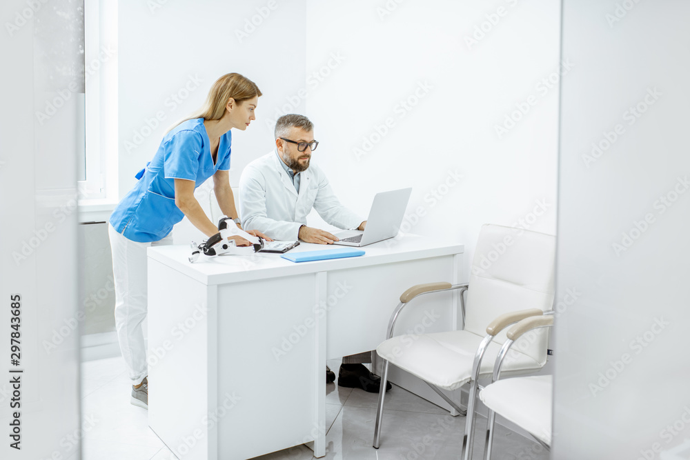 Obraz premium Senior otolaryngologist and female assistant having a medical meeting, working with laptop at the bright medical office interior