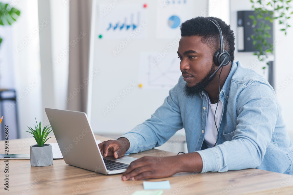 Black customer support manager with headset using laptop in office Black customer support manager with headset using laptop in office
