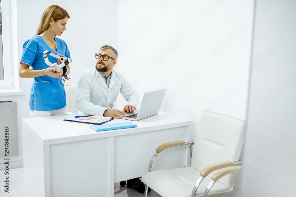 Senior otolaryngologist and female assistant having a medical meeting, working with laptop at the bright medical office interior Senior otolaryngologist and female assistant having a medical meeting, working with laptop at the bright medical office interior