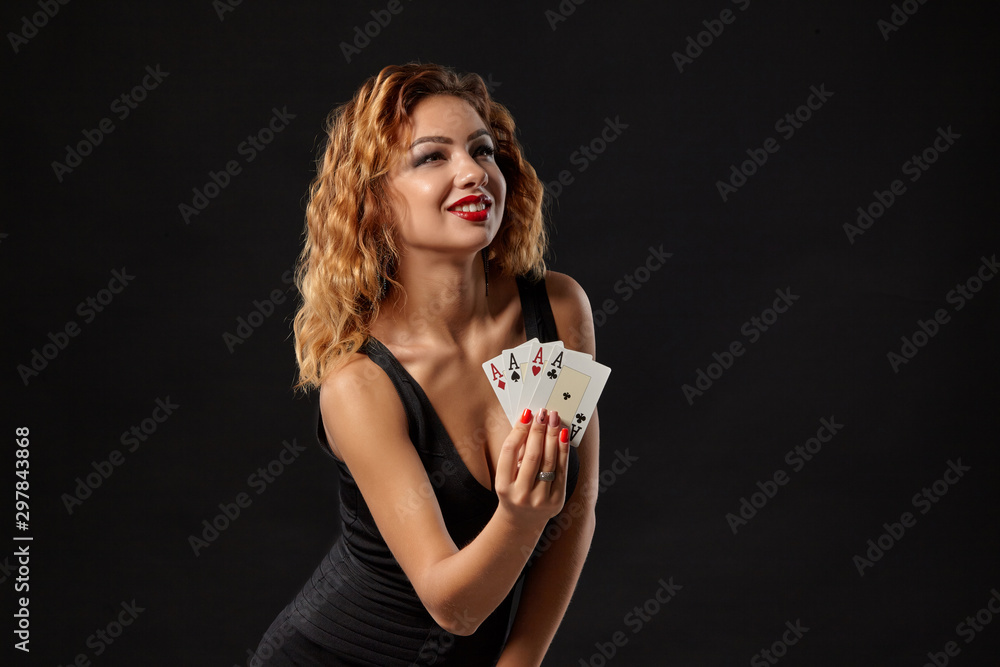 Naklejka premium Ginger girl wearing a dark dress is posing holding four aces in her hands standing against a black studio background. Casino, poker. Close-up.