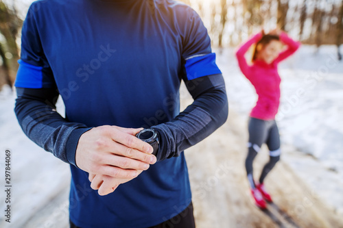Fit sporty caucasian man in sportswear checking out on smart watch heart rate after running. In background is his female friend tying hair. Wintertime. Outdoor fitness concept.