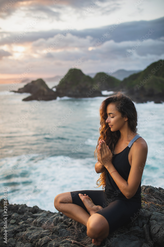 Young woman praying and meditating alone at sunset with beautiful ocean and mountain view. Self-analysis and soul-searching. Spiritual and emotional concept