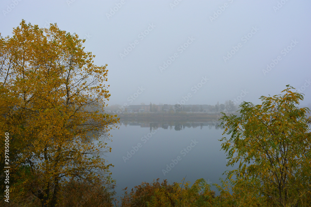 Beautiful landscape, autumnal nature with white dense fog over Career Lake, Dnipro city, Ukraine. Tall trees and large shrubs with yellow leaves around a clean and beautiful lake.