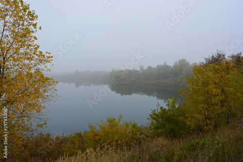 Beautiful landscape, autumnal nature with white dense fog over Career Lake, Dnipro city, Ukraine. Tall trees and large shrubs with yellow leaves around a clean and beautiful lake.