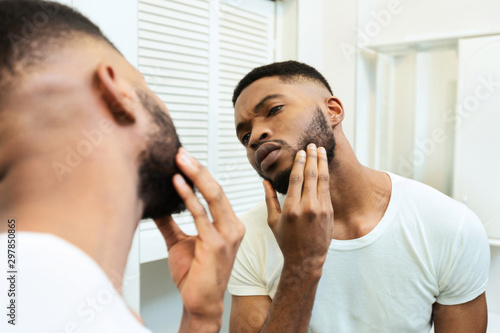 Concentrated african guy looking at his beard at bathroom mirror