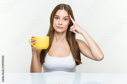 Young caucasian woman holding a cup pointing his temple with finger, thinking, focused on a task.