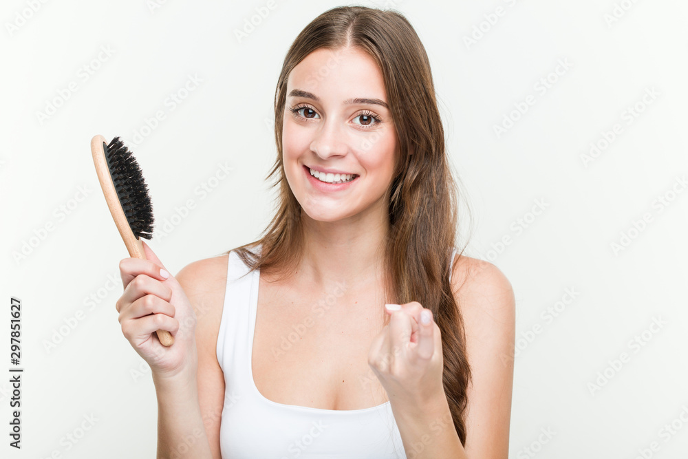 Young caucasian woman holding a hair brush pointing with finger at you as if inviting come closer. Young caucasian woman holding a hair brush pointing with finger at you as if inviting come closer.