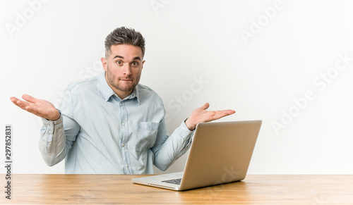 Young handsome man working with his laptop confused and doubtful shrugging shoulders to hold a copy space.