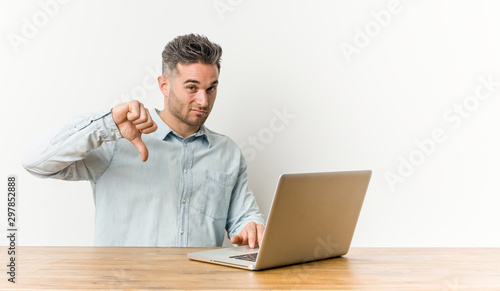 Young handsome man working with his laptop showing a dislike gesture, thumbs down. Disagreement concept.