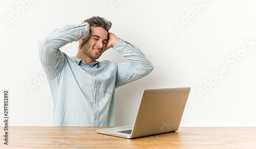 Young handsome man working with his laptop laughs joyfully keeping hands on head. Happiness concept.