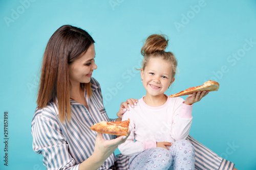 Mother and daughter eating pizza sitting on the floor in pajamas isolated over the blue background