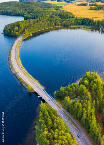 Landscape of an asphalt road. View from above on the road going along the blue lake, fields and forest. Paijanne National Park, Finland.