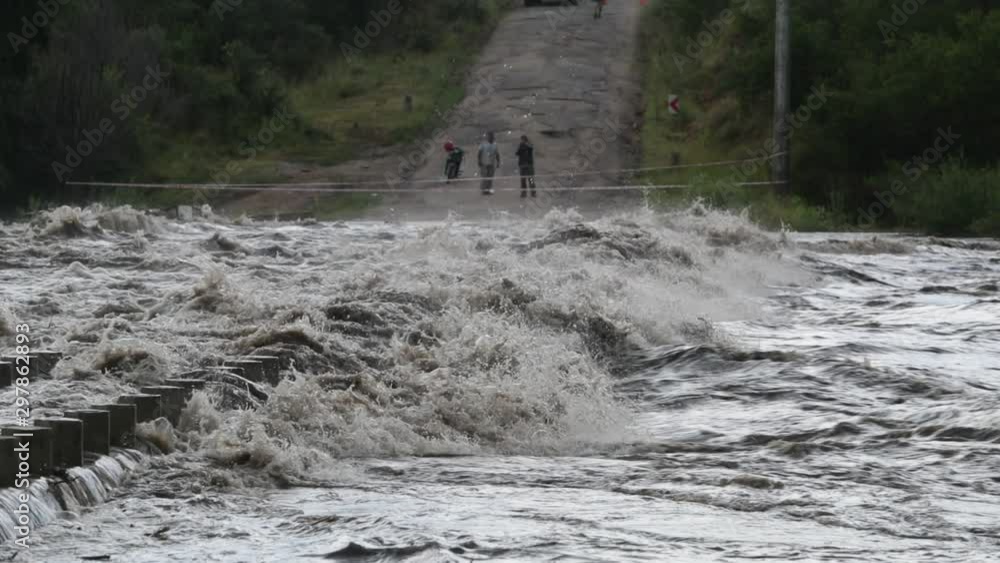 Slow motion of strong violent river passing over bridge, construction. People and fence across river at background. Flood in Mina Clavero, growing river, Cordoba, Argentina Slow motion of strong violent river passing over bridge, construction. People and fence across river at background. Flood in Mina Clavero, growing river, Cordoba, Argentina