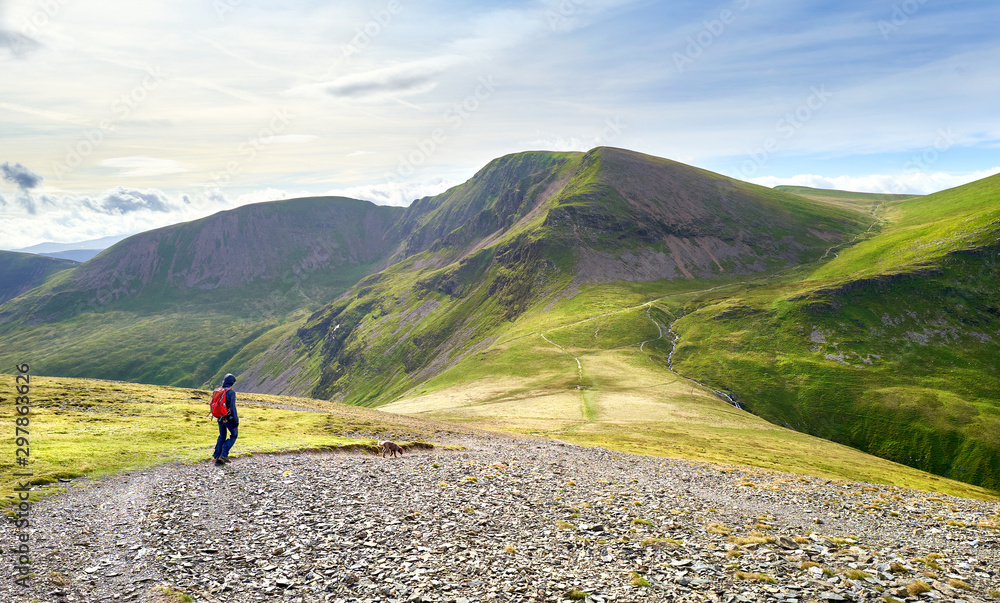 A hiker and their dog walking along a rocky path towards the summit of Sail and Crag Hill in the Lake District, England, UK.